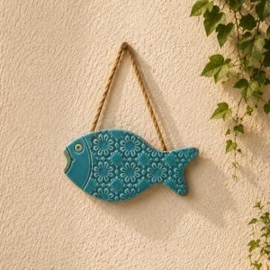 Può includere: Un pesce decorativo in ceramica color turchese con un motivo floreale in rilievo ripetuto su tutto il corpo. Il pesce presenta un occhio singolo e la bocca aperta, ed è appeso tramite una spessa corda di juta intrecciata color naturale.