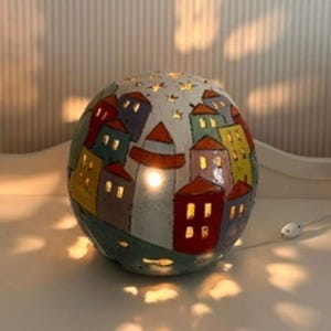 May include: A spherical ceramic lamp featuring a hand-painted village scene with colourful houses in red, yellow, blue, and green. The surface has cut-out windows and star shapes that emit light, creating a warm glow and casting patterns on surfaces.