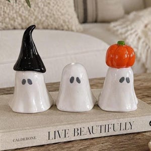 以下が含まれることがあります： 「Live Beautifully」というタイトルのベージュの本の上に置かれた、3体の白い陶器製の幽霊の置物。左の幽霊は黒いとんがり帽子をかぶり、中央の幽霊は何も身につけておらず、右の幽霊は頭に小さなオレンジ色のカボチャを乗せている。