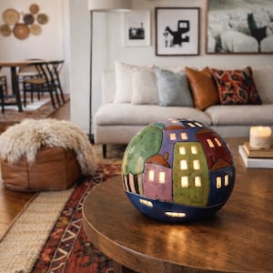 May include: A spherical ceramic luminary featuring a hand-painted village scene with colourful houses, trees, and illuminated windows. The round lamp displays a textured surface with shades of blue, green, red, and purple, casting a warm glow.