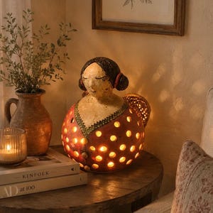 Può includere: Una lampada a forma di figura in ceramica, rotonda e cava, che raffigura una persona con capelli scuri, che indossa un abito rosso con bordo verde e ritagli circolari che emettono una luce calda. La figura ha gli occhi chiusi e un volto sereno.