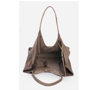 May include: A taupe suede tote bag with a long shoulder strap and a gold chain detail. The bag has a large open interior and a smaller handle. The bag is made of a soft, suede-like material.