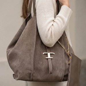 Puede incluir: Un bolso tote grande de ante color topo con un adorno metálico dorado. El bolso tiene una pequeña bolsa a juego sujeta con una cadena dorada. El bolso se lleva al hombro.