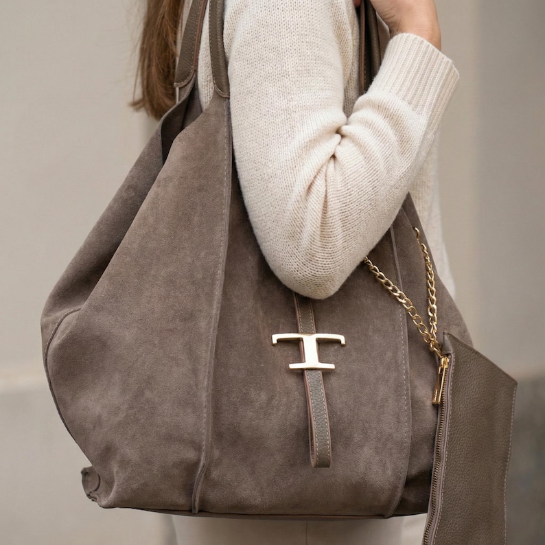 May include: A large, taupe suede tote bag with a gold-colored metal accent. The bag has a matching small pouch attached with a gold chain. The bag is carried over the shoulder.