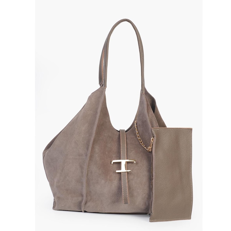 May include: A taupe suede tote bag with a matching pouch. The bag features a long handle, a gold-tone metal accent, and a delicate chain detail. The pouch is a rectangular shape and is made of a smooth leather material.