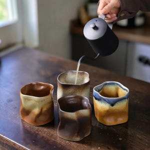 Puede incluir: Cuatro tazas de café de cerámica hechas a mano con formas orgánicas y una variedad de esmaltes en tonos tierra, incluyendo marrón, beige y azul. Una tetera de cuello de cisne negra vierte líquido en una de las tazas.