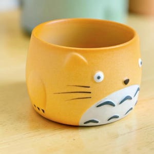 Könnte beinhalten: Orangefarbene Keramiktasse mit einem Cartoon-Katzen-Design. Die Tasse hat weiße und schwarze Details für Augen, Nase und Schnurrhaare. Die Tasse hat eine abgerundete Form und ist als Katze gestaltet.