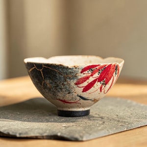 May include: A ceramic bowl with a unique design. The bowl features a white base with a blue and black section and red leaf-like patterns. The rim has a scalloped edge, and the base is black. The bowl is placed on a gray stone surface.