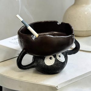 May include: A novelty ashtray with a dark brown bowl-shaped top holding a cigarette, and a black, round base with large white eyes. The ashtray has two curved handles and is on a white surface.