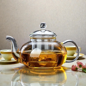 Puede incluir: Una tetera de vidrio transparente con asa y boquilla de acero inoxidable, llena de té preparado. La tetera se encuentra sobre una superficie reflectante, con dos tazas y platillos amarillos al fondo.