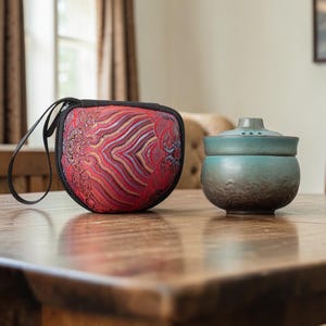 Può includere: Un astuccio con motivo rosso e oro con cerniera e manico neri è posto accanto a una piccola teiera in ceramica color verde acqua con coperchio. Gli oggetti sono su un tavolo di legno.