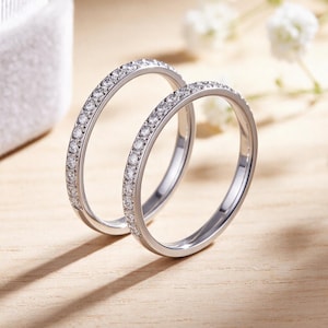 May include: Two silver-toned metal rings featuring a continuous row of small, clear, round-cut stones set along the outer band. The rings are positioned upright, showcasing a polished metallic finish and a slim, circular design with sparkling accents.