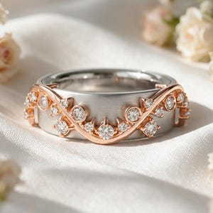 May include: A two-tone ring featuring a brushed silver-colored band overlaid with a rose gold-colored vine motif. The vine design is accented with multiple small, clear round-cut stones set along the curved, organic-shaped metal branches.