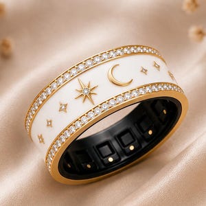 Puede incluir: Un anillo de banda ancha con una incrustación central de esmalte blanco decorada con motivos celestiales dorados, incluyendo una luna creciente y estrellas. El anillo está bordeado por dos filas de piedras claras engastadas y forro negro.