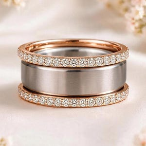 Peut inclure: Un ensemble de trois bagues composé d'un large anneau central en argent brossé encadré par deux anneaux plus fins en or rose. Chaque anneau en or rose est serti d'une rangée continue de petites pierres claires taillées en rond.