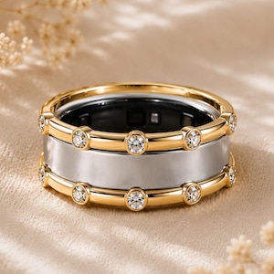 May include: A wide band ring featuring a central silver-toned metal band flanked by two gold-toned metal rails. Each gold rail is set with a series of small, clear, round-cut stones spaced evenly around the circumference of the ring.