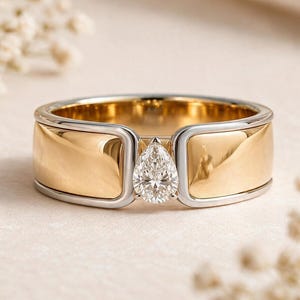 May include: A two-tone ring featuring a wide, polished yellow gold band with silver-toned edges. A single pear-shaped, clear faceted gemstone is set in the center, held by a silver-toned frame that connects the two sides of the gold band.