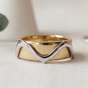May include: A two-tone ring featuring a wide, polished yellow gold band overlaid with a slender, wavy silver-toned band that forms a V-shape at the center. The design combines contrasting metallic finishes in a layered, structured composition.