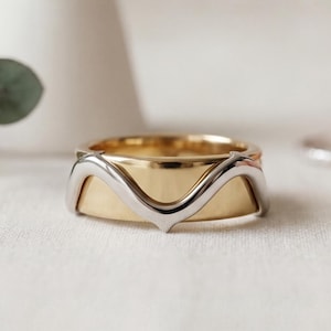 May include: A two-tone ring featuring a wide, polished yellow gold band overlaid with a slender, wavy silver-toned metal band that forms a central V-shape. The design combines a smooth, flat inner band with a contoured, elevated outer layer.