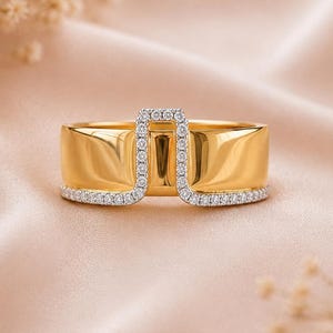 Peut inclure: Une large bague en or dotée d'un pont central en forme de U. Les bords de l'anneau et le pont en U sont incrustés de petites pierres claires taillées en rond. La bague présente une finition polie et réfléchissante ainsi qu'un anneau lisse.