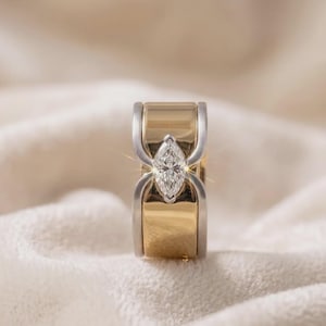 May include: A wide band ring featuring a polished yellow gold finish with silver-toned metal accents on the edges. A single marquise-cut clear gemstone is set vertically in the center, held by four prongs that extend from the silver-toned side rails.