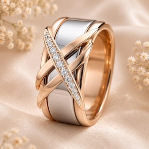May include: A wide band ring featuring a polished silver-tone centre section overlaid with rose gold-tone metal bands that cross in an X-shape. One of the crossing rose gold bands is set with a row of small, clear, round-cut sparkling stones.