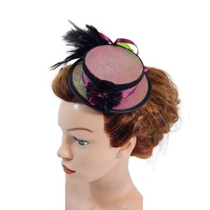 Op de afbeelding: Een kleine roze en groene hoge hoed met een zwarte veer en een paarse bloemaccent. De hoed wordt gedragen op een mannequinhoofd met bruin haar.