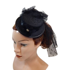 Op de afbeelding: Een zwarte mini hoge hoed met een gestructureerd damastpatroon, een zwarte satijnen lintband en een decoratieve zilverkleurige broche met een heldere steen in het midden. De hoed is versierd met zwart tule gaas dat aan de zijkant hangt.