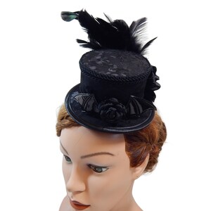 Op de afbeelding: Mini zwarte hoge hoed met een vleermuisvleugelontwerp en een zwarte roos. De hoed is versierd met zwarte veren en een groene accentkleur. Een gothic accessoire.