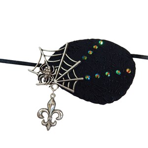 Pode incluir: Atadura de olho preta com uma teia de aranha e um amuleto de aranha, decorado com pedras de cristal verde iridescentes. O amuleto tem um pingente de flor-de-lis.