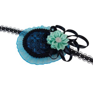 Op de afbeelding: Een decoratief ooglapje met een zwart kanten bandje. De patch heeft een blauw en zwart ovaal ontwerp met een turquoise bloem en zwarte lintaccenten. De bloem heeft een strass steentje in het midden.