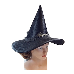 Op de afbeelding: Zwarte heksenhoed met een brede rand en een hoge, puntige kroon. De hoed is versierd met een zilveren vleermuisembleem en kanten afwerking. Een Halloween kostuumaccessoire.