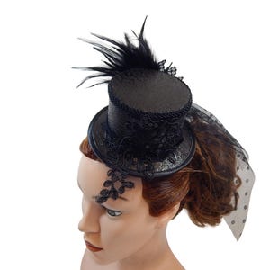 Op de afbeelding: Een zwarte mini hoge hoed fascinator met ingewikkelde zwarte kanten bloemapplicaties, een getextureerde kroon en een decoratieve zwarte veer. Het accessoire bevat een doorschijnende zwarte sluier met stippen aan de zijkant.