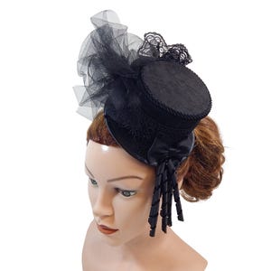 Op de afbeelding: Een zwarte mini-hoge hoed fascinator met een platte ronde kroon met gevlochten rand, een brede rand en decoratief zwart tule net. Het accessoire is versierd met gedetailleerd zwart kant, een kleine satijnen strik en hangende lintkrullen.