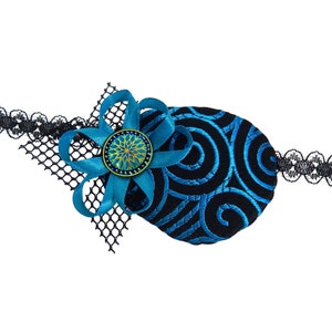 Pode incluir: Um tapa-olho azul e preto com um padrão de espiral e uma flor azul com um centro dourado. O tapa-olho está preso a uma faixa de renda preta.