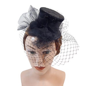 Op de afbeelding: Een zwarte mini hoge hoed fascinator met een gestructureerde cilindrische kroon en een platte rand. De hoed is versierd met gelaagde zwarte tule en een delicate sluier die over de voorkant valt, wat zorgt voor een getextureerd uiterlijk.