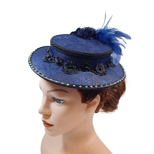 Op de afbeelding: Een marineblauwe hoed met een kleine rand en een cilindrische kroon. De hoed is versierd met zwarte bloemaccenten, een zwarte touwrand en blauwe veren. Strassstenen omzomen de rand.
