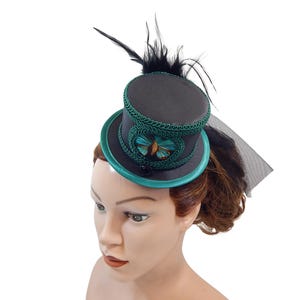 Op de afbeelding: Een kleine zwarte hoge hoed fascinator met een groenblauwe rand en afwerking, versierd met een centrale vlinder, zwarte veren en een doorschijnende zwarte sluier die vanaf de achterkant van de kroon naar beneden valt, op een witte achtergrond.