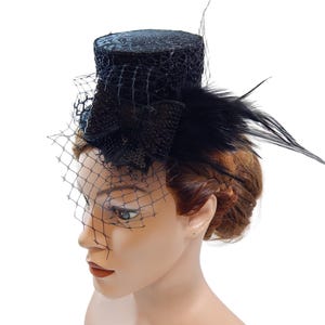 Op de afbeelding: Een zwarte fascinator hoed met een kleine, cilindrische kroon en een delicate netsluier. De hoed is versierd met zwarte veren en pailletten, wat een vintage of formele uitstraling creëert. De hoed is ontworpen om op het hoofd te worden gedragen.