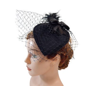 May include: A black heart-shaped hat with a delicate net veil. The hat features a bow and feather detail. The hat is a vintage style accessory.
