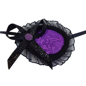 May include: A purple eye patch with black lace trim and a black bow with a sparkly purple button. The eye patch is attached to a black ribbon.