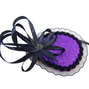 May include: A decorative eye patch featuring a purple oval base with a floral pattern, trimmed with black lace. A black ribbon and bow with a silver-colored center embellish the patch. The accessory is designed for costume or fashion use.