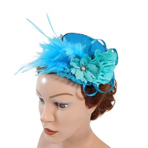 Op de afbeelding: Een turquoise fascinator met veren en bloemaccenten. De hoofdtooi heeft een kleine, ronde basis versierd met veren, een stoffen bloem en decoratieve linten. Een parelachtige kraal bevindt zich in het midden van de bloem.