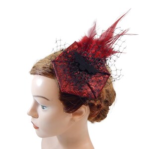 May include: A red and black decorative hair clip with a bat-shaped embellishment. The clip features a square base with a textured pattern, adorned with red feathers and a black net veil. The accessory is designed for formal events.