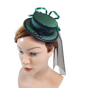 Op de afbeelding: Een kleine groene hoge hoed met zwarte bies en een zwarte sluier. De hoed heeft een patroon en wordt gedragen op een mannequinhoofd.