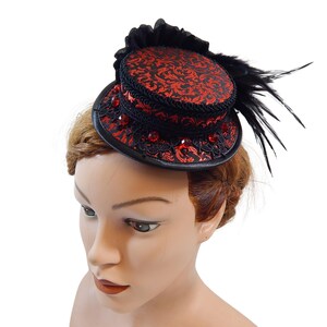 Op de afbeelding: Een kleine zwarte en rode hoge hoed met een bloemmotief. De hoed heeft rode juwelen en zwarte veren. Deze opvallende hoed is een geweldig accessoire voor thema-evenementen en feesten.