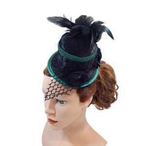 Op de afbeelding: Een zwarte fluwelen mini-hoge hoed met een felgroene satijnen rand, een decoratieve groene gevlochten bies rond de kroon, zwarte veren en een zwarte netsluier die over de voorkant van de hoed valt, wat zorgt voor een gelaagd textuurbeeld.