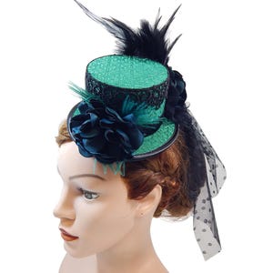 Op de afbeelding: Een teal en zwarte mini-cilinderhoed met een bloemaccent en veren. De hoed heeft een getextureerde teal kroon met zwarte kanten rand, een grote donkerblauwe bloem en een zwarte sluier met witte stippen. Een stijlvol accessoire.