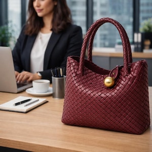 May include: A burgundy leather handbag with a woven design, featuring two braided handles and a gold-colored clasp. The bag is sitting on a wooden desk, with a woman working on a laptop in the background.