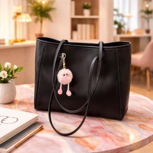 May include: A black tote bag with long handles, on a pink marble table. A pink fluffy keychain with a face and legs hangs from the handle. A small white flower pot and a book are in the foreground.